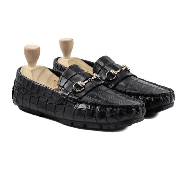 May include: A pair of black leather loafers with a crocodile texture. The shoes have a gold-colored metal accent across the top and are displayed with wooden shoe trees. The loafers are designed for a classic, sophisticated look.