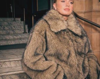 May include: A faux fur coat in shades of brown and tan. The coat has a wide collar and large sleeves. The person is standing in front of a marble staircase and a dark doorway.