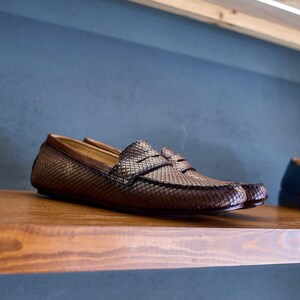 May include: A pair of brown loafers featuring a textured, diamond-patterned surface across the upper. The shoes have a classic slip-on design with a horizontal strap across the vamp and a smooth brown leather trim along the collar and heel.