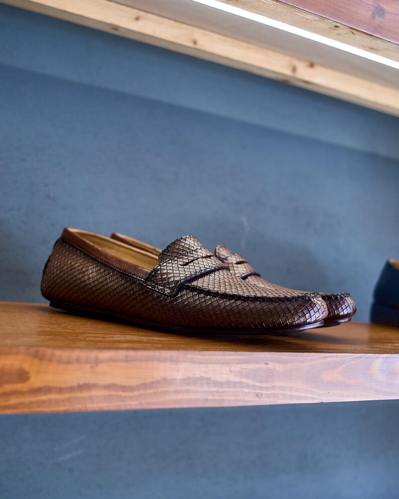 May include: A pair of brown loafers featuring a textured, diamond-patterned surface across the upper. The shoes have a classic slip-on design with a horizontal strap across the vamp and a smooth brown leather trim along the collar and heel.