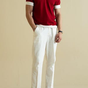 Op de afbeelding: Een persoon draagt een rood poloshirt met witte kraag en manchetten, gecombineerd met een witte linnen broek en bruine schoenen. De outfit is een klassieke, casual stijl.