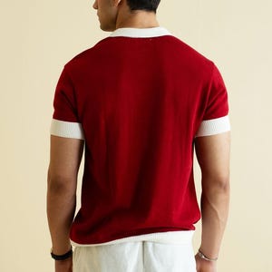Op de afbeelding: Een rood shirt met korte mouwen en witte details op de kraag en mouwen. De persoon draagt een witte broek. Het shirt is effen van kleur en lijkt van een zacht materiaal te zijn.