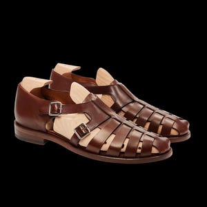 May include: A pair of brown leather sandals with a cage-like design. The sandals have a buckle closure and a wooden shoe form inside. The shoes are set against a black background.