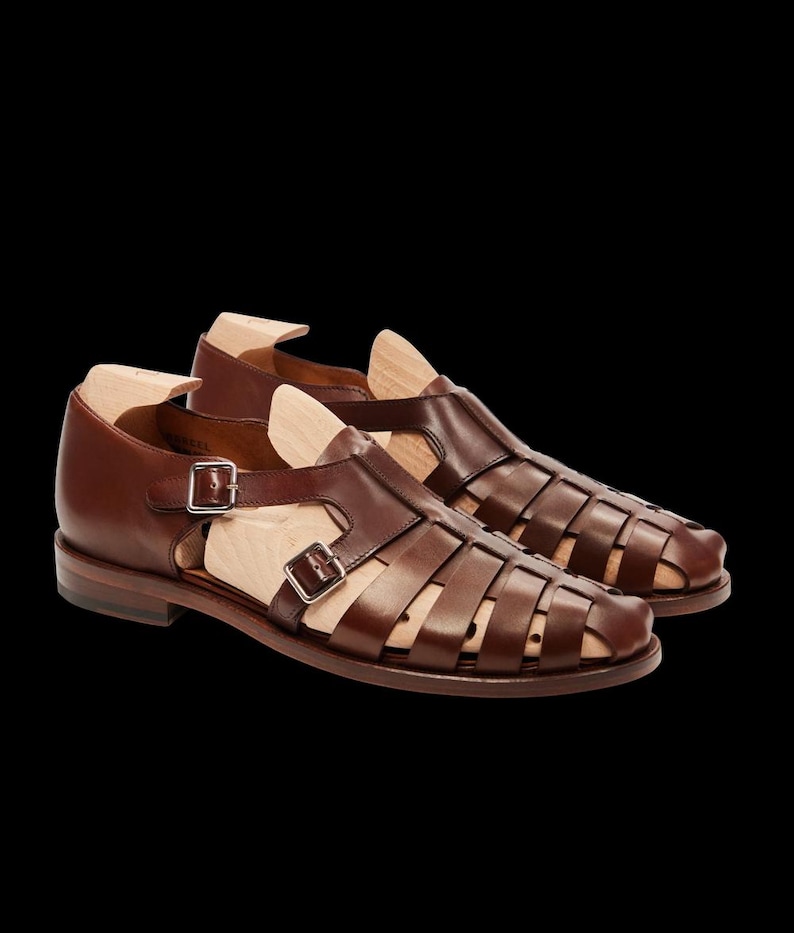 May include: A pair of brown leather sandals with a cage-like design. The sandals have a buckle closure and a wooden shoe form inside. The shoes are set against a black background.