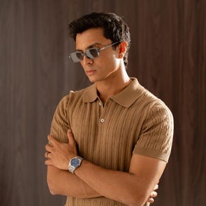 May include: A tan cable knit polo shirt with short sleeves and a button-down collar. The shirt is paired with blue jeans and a silver watch with a blue face. The person is wearing rectangular sunglasses.