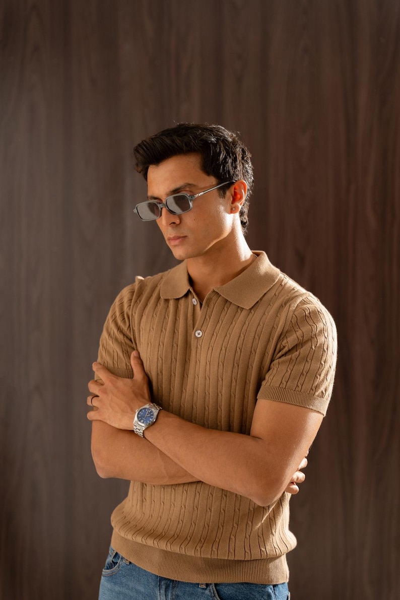 May include: A tan cable knit polo shirt with short sleeves and a button-down collar. The shirt is paired with blue jeans and a silver watch with a blue face. The person is wearing rectangular sunglasses.