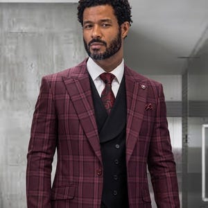 May include: A burgundy plaid blazer worn over a black vest and a white collared shirt. The ensemble includes a dark red patterned necktie and a matching pocket square. The blazer features wide lapels and a single button closure at the front.