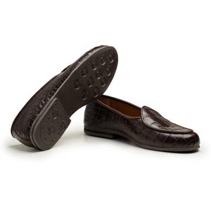 Puede incluir: Un par de mocasines de cuero marr&oacute;n con textura de piel de cocodrilo. Un zapato est&aacute; en posici&oacute;n vertical y el otro est&aacute; volteado, revelando la suela. Los zapatos tienen un dise&ntilde;o cl&aacute;sico y un acabado pulido.