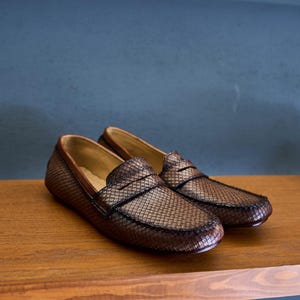 May include: A pair of brown leather loafers featuring a textured, woven-like grid pattern across the upper surface. The shoes have a classic penny loafer strap detail across the vamp and a smooth, light tan interior lining visible at the opening.