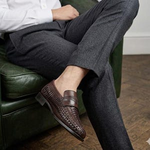 May include: Brown leather loafers with a woven design. The shoes have a classic penny loafer style with a low heel. The image shows the shoes on a person sitting down, wearing gray pants and a white shirt.
