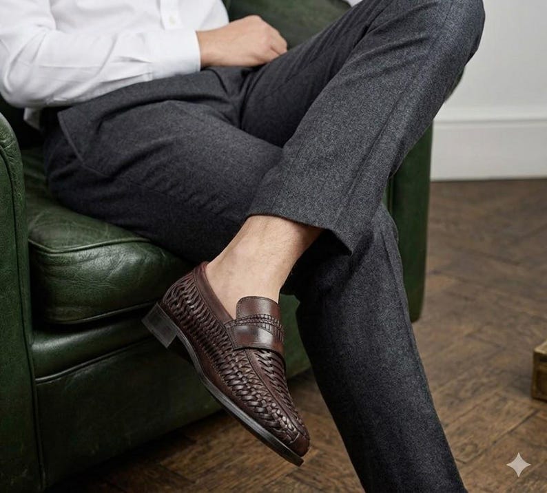 May include: Brown leather loafers with a woven design. The shoes have a classic penny loafer style with a low heel. The image shows the shoes on a person sitting down, wearing gray pants and a white shirt.