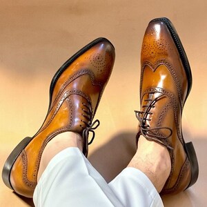 Puede incluir: Un par de zapatos brogue de cuero marr&oacute;n con intrincadas perforaciones y un dise&ntilde;o cl&aacute;sico con cordones. Los zapatos tienen un acabado pulido y una suela marr&oacute;n oscuro. Los zapatos se muestran sobre un fondo neutro.