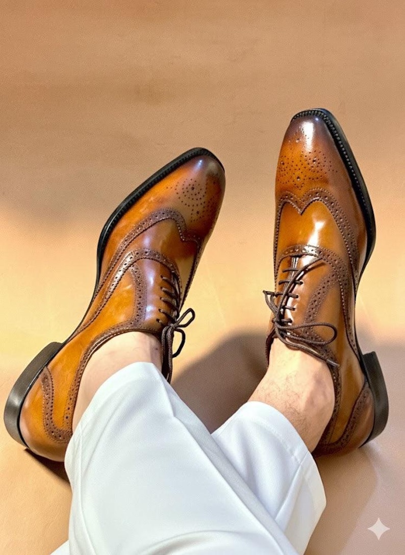 Puede incluir: Un par de zapatos brogue de cuero marr&oacute;n con intrincadas perforaciones y un dise&ntilde;o cl&aacute;sico con cordones. Los zapatos tienen un acabado pulido y una suela marr&oacute;n oscuro. Los zapatos se muestran sobre un fondo neutro.