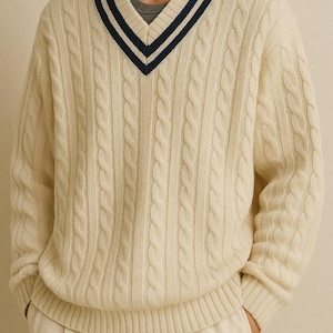 May include: Cream-colored cable knit sweater with a navy and white striped V-neck. The sweater has a ribbed hem and cuffs. The model is wearing a gray undershirt and cream-colored pants. The sweater is a classic style.