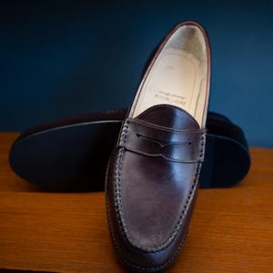 May include: A pair of dark brown leather penny loafers. The shoes have a classic design with a rounded toe, a strap across the vamp, and visible stitching. The soles are dark, and the shoes are displayed on a wooden surface.