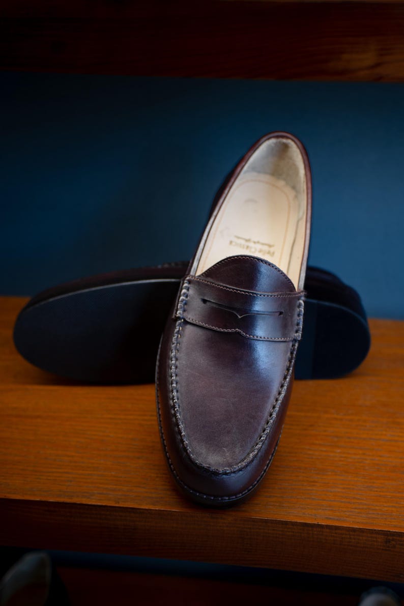 May include: A pair of dark brown leather penny loafers. The shoes have a classic design with a rounded toe, a strap across the vamp, and visible stitching. The soles are dark, and the shoes are displayed on a wooden surface.