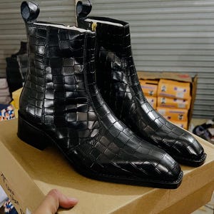 May include: A pair of black ankle boots with a faux crocodile skin texture. The boots have a square toe, a small heel, and a zipper closure. The boots are sitting on a cardboard box.