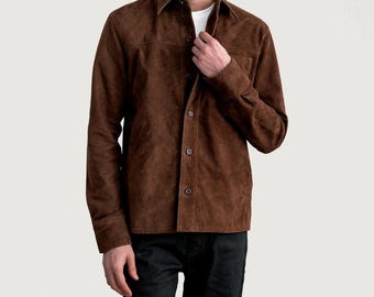 May include: A brown suede button-up shirt with a classic collar and long sleeves. The shirt has a chest pocket and is worn over a white t-shirt. The model is wearing black jeans.