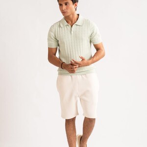 May include: A pale green polo shirt with a ribbed design and short sleeves, worn with cream shorts and tan loafers. The outfit is set against a white background, creating a clean, minimalist aesthetic.