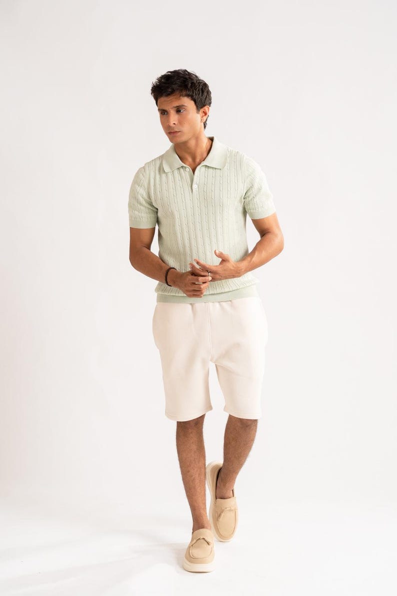 May include: A pale green polo shirt with a ribbed design and short sleeves, worn with cream shorts and tan loafers. The outfit is set against a white background, creating a clean, minimalist aesthetic.