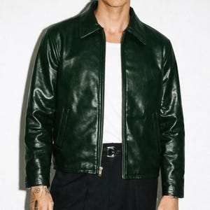 Op de afbeelding: Een donkergroene leren jas met een kraag en een ritssluiting over de hele lengte. De jas heeft twee voorzakken en een zwarte riem met een zilveren gesp. Het model draagt er een wit t-shirt onder.