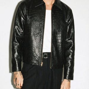 May include: A black leather jacket with a textured finish and a zip fastening. The jacket features a traditional collar and a subtle embossed pattern. The model is wearing a white t-shirt and black trousers. The jacket's design resembles crocodile skin.