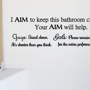 Könnte beinhalten: Eine weiße Badezimmerwand mit einem schwarzen Vinyl-Aufkleber, auf dem steht: "I AIM to keep this bathroom clean. Your AIM will help. Guys: Stand closer. It's shorter than you think. Gals: Please remain seated for the entire performance."