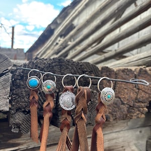 May include: A collection of five keychains with silver-toned metal accents and brown leather braids. Each keychain features a different decorative element, including turquoise and patterned silver designs. The keychains are displayed against a rustic wooden background.