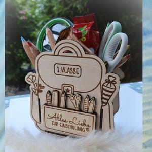 Könnte beinhalten: Schreibtisch-Organizer in Rucksackform aus Holz, gefüllt mit Bleistiften, Schere und einem Snack. Der Organizer trägt die Gravuren "1. Klasse" und "Alles Liebe zur Einschulung". Ein charmantes Geschenk für einen Schüler.