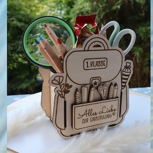 May include: A wooden desk tidy in the shape of a school backpack, containing pencils, scissors, and a magnifying glass. The organizer has the text "1. Klasse" and "Alles Liebe zur Einschulung 83" engraved on it.