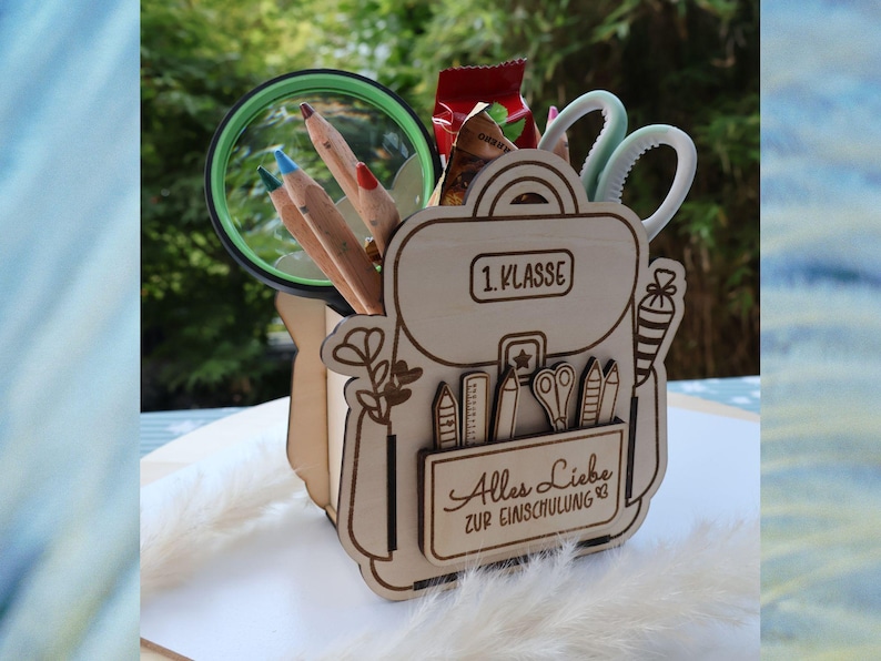 May include: A wooden desk tidy in the shape of a school backpack, containing pencils, scissors, and a magnifying glass. The organizer has the text "1. Klasse" and "Alles Liebe zur Einschulung 83" engraved on it.