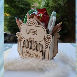 May include: Wooden desk tidy in the shape of a backpack, filled with pencils, scissors, and other stationery. The organiser has the text "1. Klasse" and "Alles Liebe zur Einschulung" engraved on it. A red snack packet is visible.