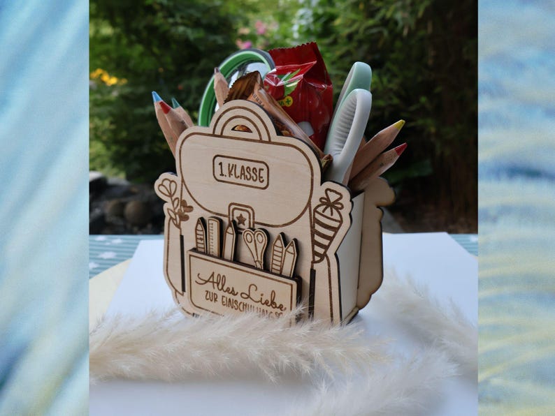 May include: Wooden desk tidy in the shape of a backpack, filled with pencils, scissors, and other stationery. The organiser has the text "1. Klasse" and "Alles Liebe zur Einschulung" engraved on it. A red snack packet is visible.
