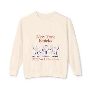 May include: A cream-colored long-sleeve sweatshirt featuring the red text New York Knicks above a blue graphic of three anthropomorphic basketball characters, with the red text 2026 NBA Champions printed below the characters on the front.