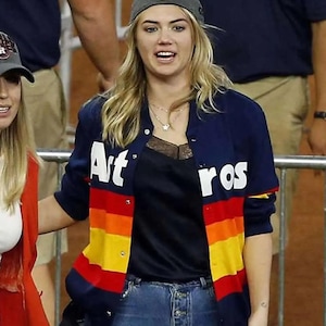 May include: A navy blue baseball-style jacket with the word "Astros" in white lettering. The jacket features a rainbow stripe design in red, orange, yellow, and a black top. The person is wearing a gray baseball cap and blue jeans.