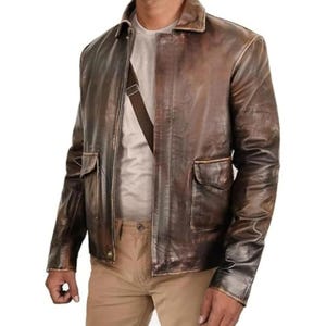Puede incluir: Chaqueta de cuero marrón con acabado envejecido. La chaqueta tiene dos bolsillos delanteros, cuello y cierre de botones. Se lleva sobre una camiseta gris claro y pantalones color beige. Se lleva un bolso bandolera marrón cruzado.