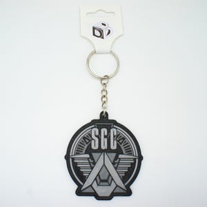 May include: A black and grey circular keyring featuring the letters SGC in a bold font above a stylised geometric emblem with wing-like details. The keyring is attached to a silver metal ring and chain, connected to a white display card with a logo.