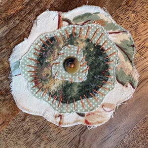 May include: A layered fabric flower applique featuring a base of cream floral patterned fabric, a middle layer of sage green polka dot fabric, and a dark green center, all secured with orange hand-stitched embroidery and a central brown button.