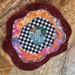 May include: A layered fabric applique featuring a dark maroon base, an orange and pink mottled layer, a black and white gingham circle, and a central green floral patch. The layers are joined with visible blue and red hand-stitched embroidery.