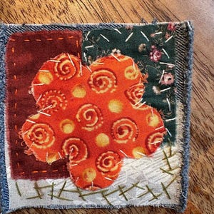May include: A square fabric patch featuring a central orange flower shape with yellow spiral patterns, layered over a dark red square, a dark green floral print square, and a white square, all joined with visible white and green hand-stitched embroidery.