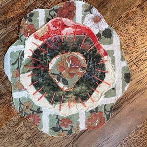 May include: A layered fabric applique featuring three circular pieces of textile stacked and stitched together. The base layer has a green and white striped floral pattern, topped with a dark green textured circle and a small central floral patch.