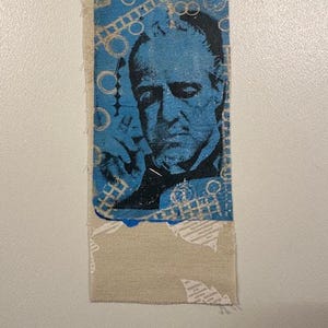 May include: A rectangular fabric patch featuring a blue and black portrait of a man. The background is blue with white geometric shapes. The bottom portion of the patch is a beige color with a white pattern.