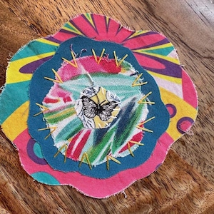 May include: A layered fabric applique featuring a circular base with a yellow, pink, and teal sunburst pattern, a solid teal middle layer, and a central white patch with a black butterfly print, all secured with yellow decorative embroidery stitches.