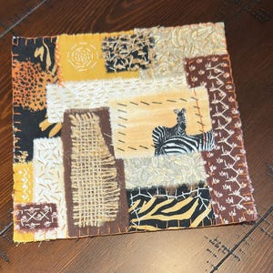 May include: A square fabric collage featuring various patches in shades of yellow, brown, black, and cream. The piece includes textured burlap, animal print patterns, and a central patch depicting two zebras, all joined by visible orange stitching.