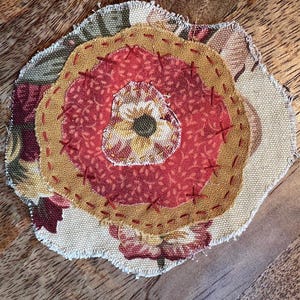 May include: A circular fabric applique featuring layered floral patterns in shades of red, gold, and cream. The piece is constructed with raw, frayed edges and secured with visible dark red cross-stitching that connects the various textile layers.