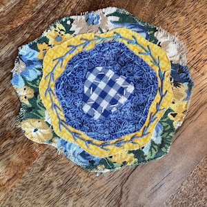 May include: A circular fabric coaster featuring layered textile scraps with frayed edges. The design includes a floral print base in green and blue, a yellow fabric ring with blue embroidery, a dark blue patterned center, and a blue gingham patch.
