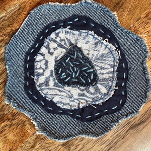 May include: A circular fabric patch featuring layered denim and patterned textile circles with frayed edges. The center is a dark blue triangle with light blue hand-stitched details, surrounded by a light blue floral print and dark blue denim rings.