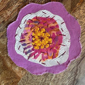 May include: A circular fabric applique featuring layered textile pieces in shades of purple, white, pink, and yellow. The center has a yellow checkered pattern, surrounded by pink and orange abstract shapes, white textured fabric, and a purple border.