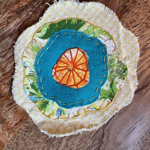 May include: A circular fabric applique featuring layered textile pieces with raw, frayed edges. The design includes a yellow and white gingham base, a green floral layer, a solid teal center, and an orange embroidered citrus slice motif.