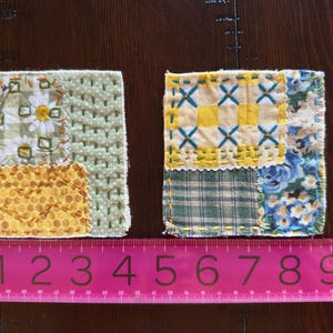 May include: Two handmade fabric coasters, each approximately 4 inches square. One features a daisy print, the other a yellow and blue patchwork design. Both are stitched with contrasting thread and have a homespun aesthetic.